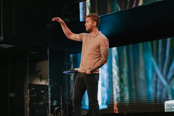 Joshua Shaw preaching on stage in a tan polo shirt with one arm extended, in front of a screen showing a muted forest scene.