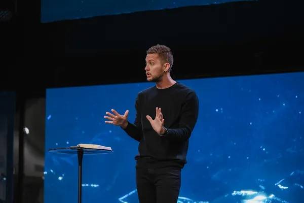 Joshua Shaw preaching in front of a blue screen, wearing a black sweater, with an open Bible on a lectern beside him.