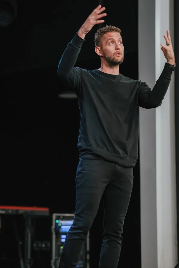 Joshua Shaw preaching on stage in a black sweater with both hands raised, against a dark background.