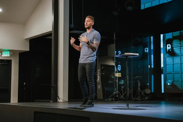 Joshua Shaw preaching on a dark stage in a grey polo shirt, gesturing with one hand beside a lectern with an open Bible.